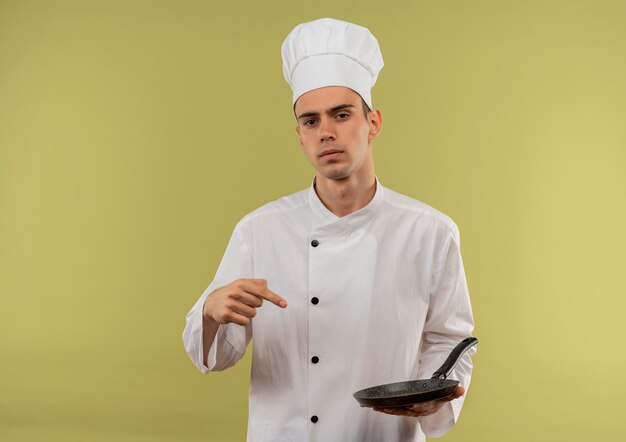 Confident young male cook wearing chef uniform points finger to frying pan in his hand on isolated green wall with copy space