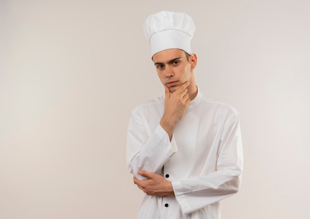 Confident young male cook wearing chef uniform putting hand on his chin on isolated white wall with copy space