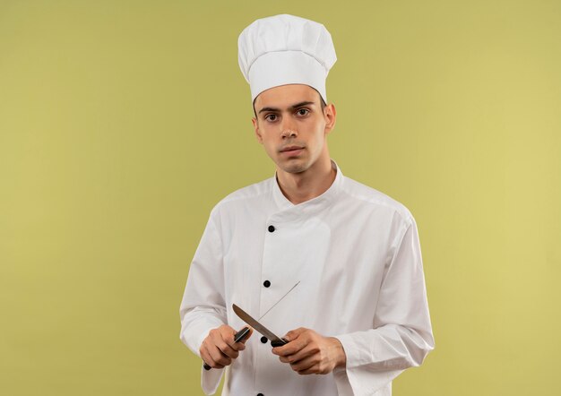 Confident young male cook wearing chef uniform sharpes a knife on isolated green wall