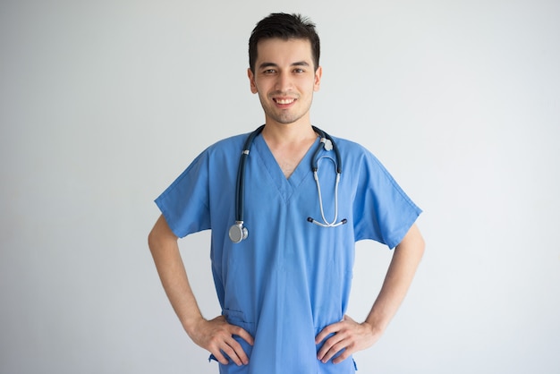 Giovane medico maschio sicuro che si leva in piedi e che mantiene le mani sulle anche. concetto di assistenza sanitaria.