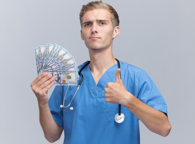 Confident young male doctor wearing doctor uniform with stethoscope holding cash showing thumb up isolated on white wall