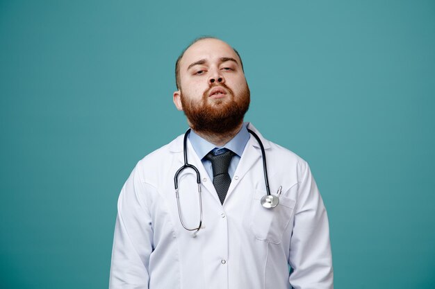 Confident young male doctor wearing medical coat and stethoscope around his neck looking at camera isolated on blue background