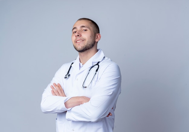 Confident young male doctor wearing medical robe and stethoscope around his neck standing with closed posture and smiling isolated on white  with copy space