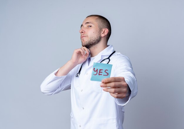 Confident young male doctor wearing medical robe and stethoscope around his neck stretching out yes note towards camera and touching his chin isolated on white  with copy space