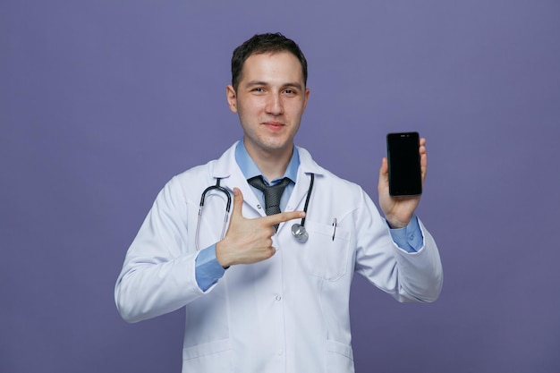 首の周りに医療ローブと聴診器を身に着けている自信を持って若い男性医師は、紫色の背景に分離されたカメラを見てそれを指している携帯電話を示しています