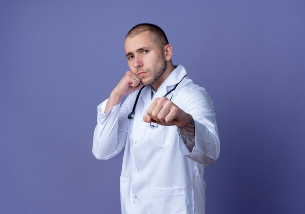 Уверенный молодой мужчина-врач в медицинском халате и стетоскопе делает боксерский жест, изолированный на фиолетовом, с копией пространства