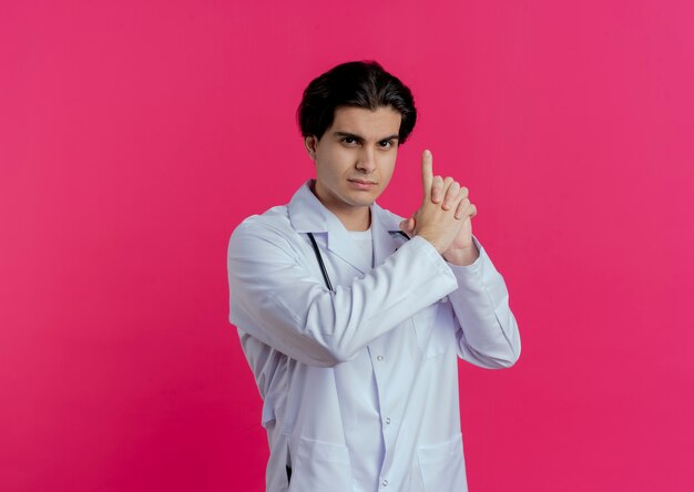 Confident young male doctor wearing medical robe and stethoscope  doing pistol gesture isolated on pink wall with copy space