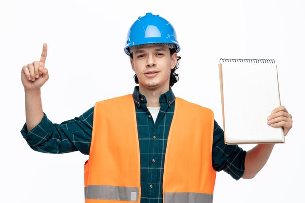 Confident young male engineer wearing safety helmet and safety vest showing note pad to camera looking at camera pointing up isolated on white background