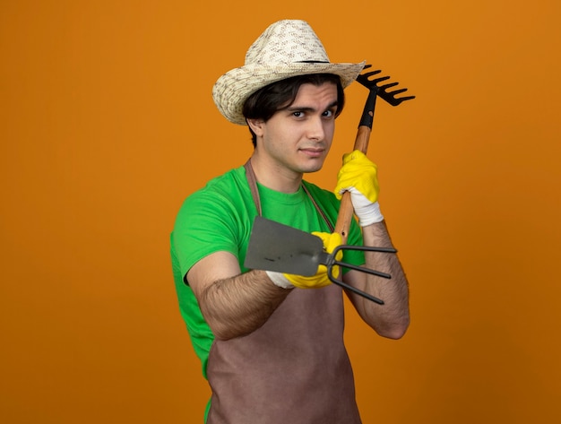 Бесплатное фото Уверенный молодой мужчина-садовник в униформе в садовой шляпе с перчатками, держащими грабли на оранжевом фоне