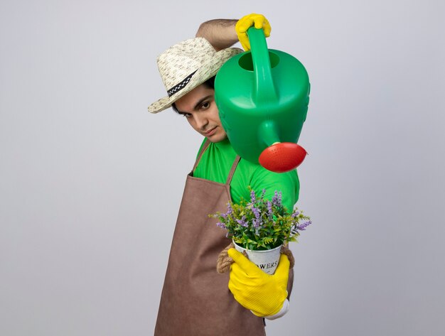 물을 수있는 화분에 꽃을 물을 원예 모자와 장갑을 착용하는 제복을 입은 자신감이 젊은 남성 정원사