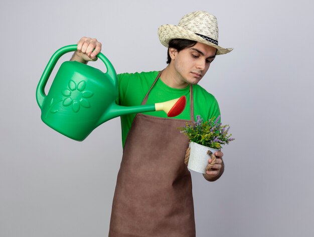 Confident young male gardener in uniform wearing gardening hat watering flower in flowerpot with watering can