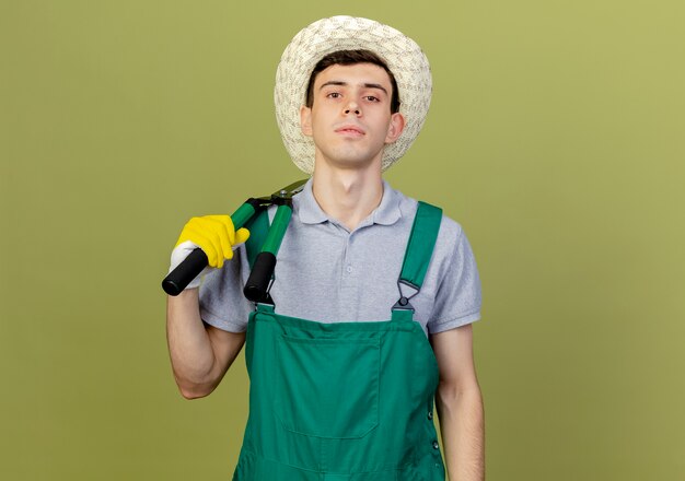 Уверенный молодой мужчина-садовник в садовой шляпе и перчатках держит ножницы на плече
