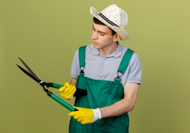 Fiducioso giovane giardiniere maschio che indossa cappello e guanti da giardinaggio tiene e guarda i tagliaunghie