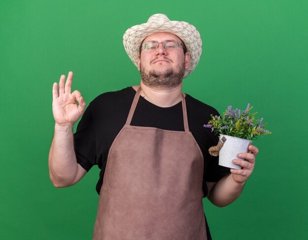 Confident young male gardener wearing gardening hat holding flower in flowerpot showing okay gesture isolated on green wall