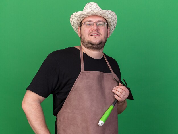 Confident young male gardener wearing gardening hat holding hoe rake putting hand on hip isolated on green wall with copy space