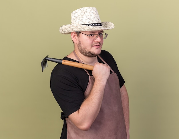 Foto gratuita fiducioso giovane giardiniere maschio che indossa cappello da giardinaggio tenendo il rastrello sulla spalla isolato sulla parete verde oliva