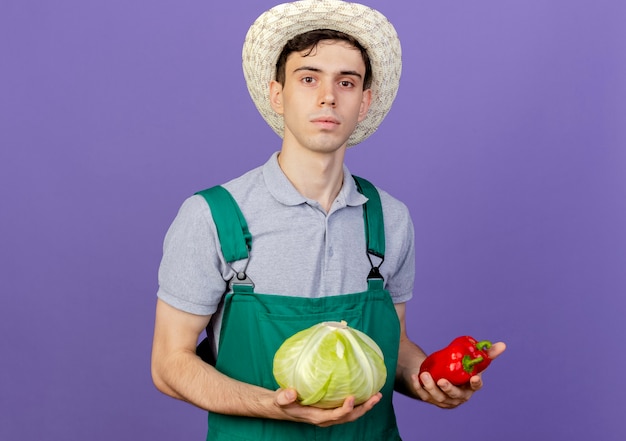 Confident young male gardener wearing gardening hat holds cabbage and peppers 