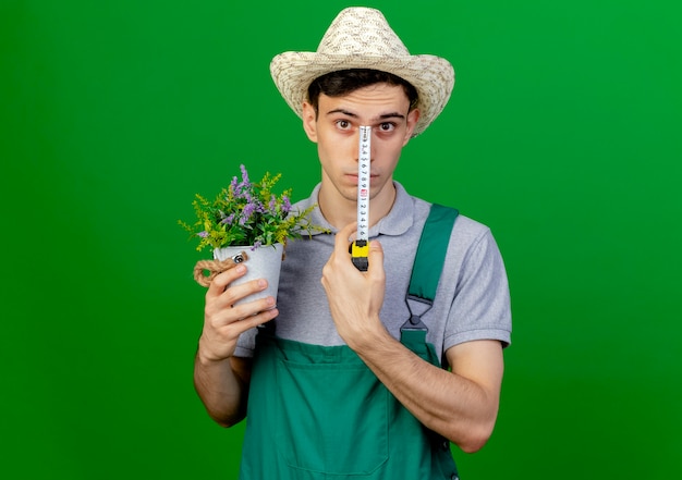 Confident young male gardener wearing gardening hat holds flowerpot and tape measure