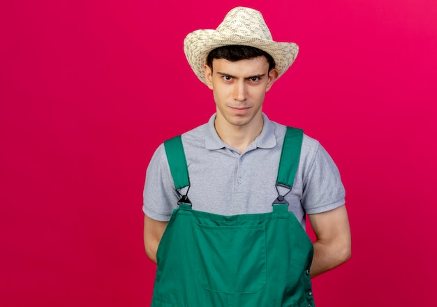 Confident young male gardener wearing gardening hat holds hands behind