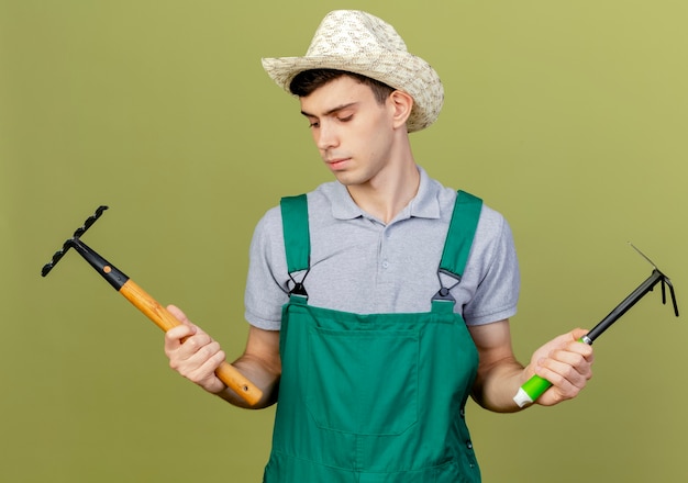 Confident young male gardener wearing gardening hat holds hoe rake and looks at rake