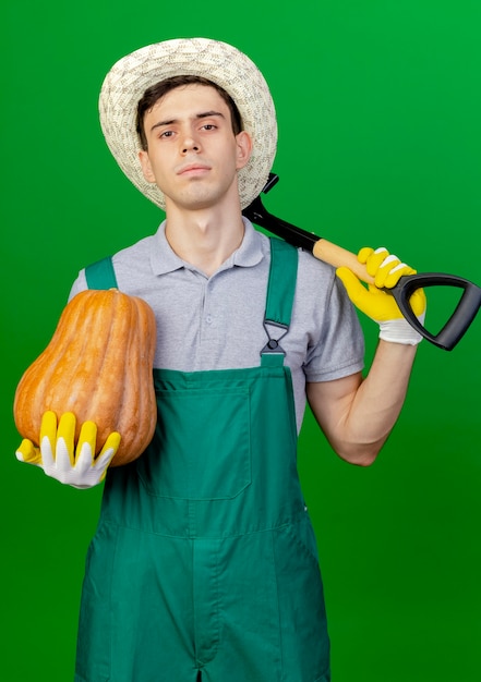 Бесплатное фото Уверенный молодой мужчина-садовник в садовой шляпе держит тыкву и лопату, изолированные на зеленом фоне с копией пространства