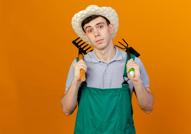 Confident young male gardener wearing gardening hat holds rake and hoe rake on shoulders