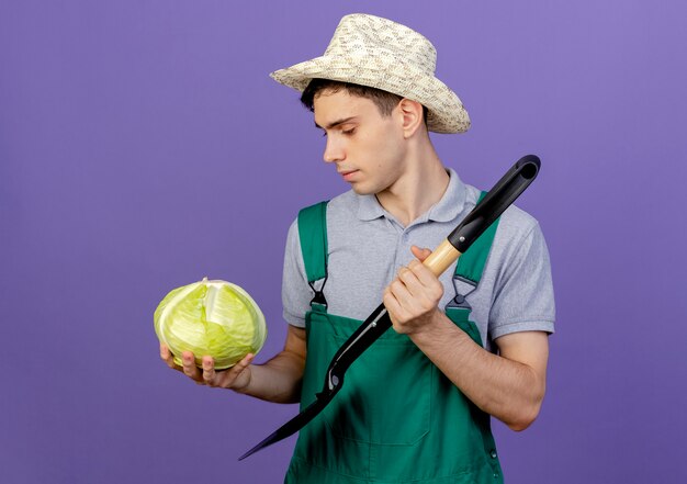 Confident young male gardener wearing gardening hat holds spade and cabbage