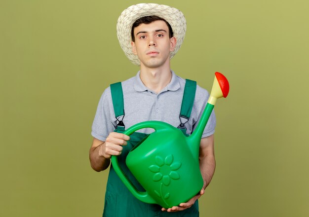 Confident young male gardener wearing gardening hat holds watering can 