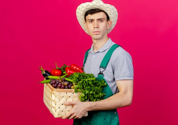 ガーデニング帽子をかぶって自信を持って若い男性の庭師は、野菜のバスケットを持って横に立っています