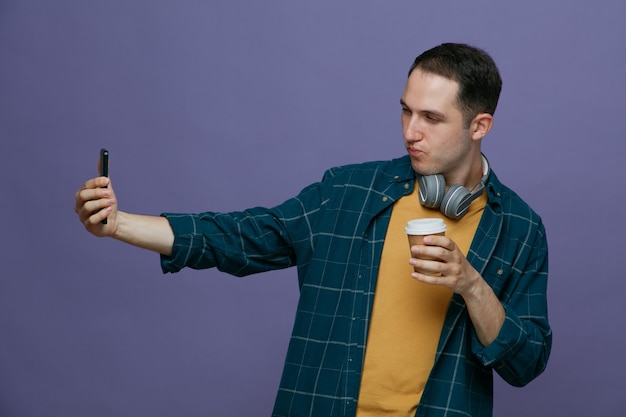 Уверенный молодой студент в наушниках на шее держит бумажную чашку кофе, растягивая мобильный телефон, делая селфи на фиолетовом фоне