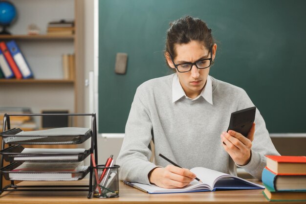 교실에 학교 도구가 있는 책상에 앉아 있는 노트북에 계산기를 들고 있는 자신감 있는 젊은 남성 교사
