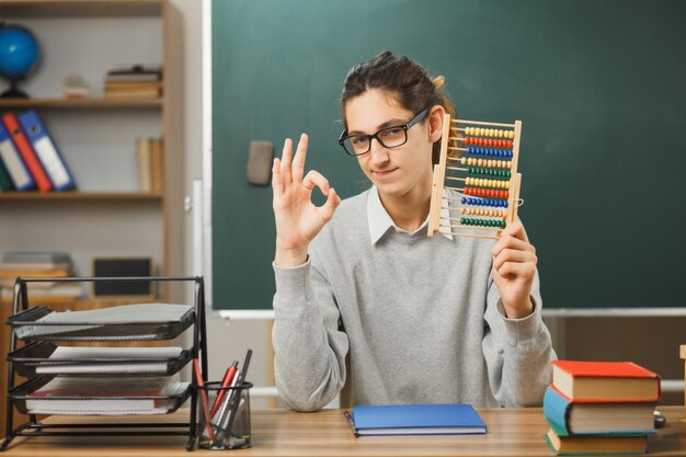 自信を持って若い男性教師がそろばんを持って黒板を指さし、教室で学校の道具を使って机に座って大丈夫なジェスチャーを示しています