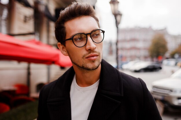 Confident young man in full suit looking away while standing outdoors in european city. Wearing eyewear.