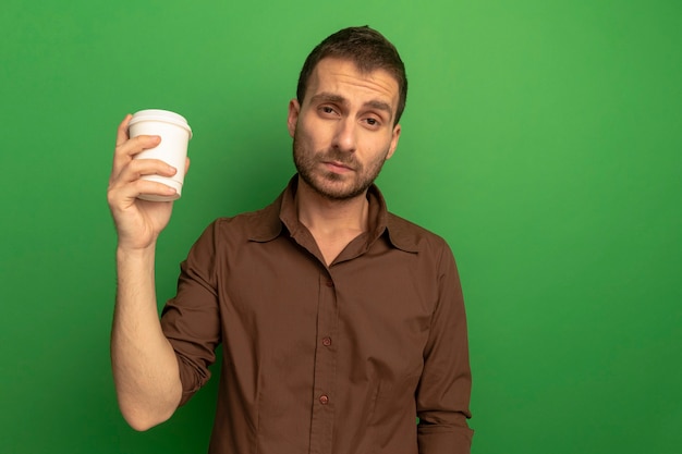 Confident young man holding plastic coffee cup looking at front isolated on green wall
