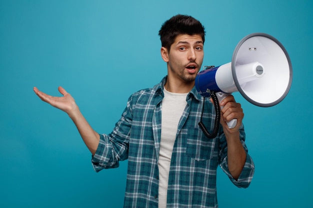 Free photo confident young man looking at camera talking into speaker showing empty hand isolated on blue background