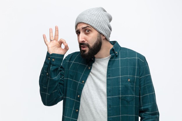Confident young man wearing winter hat looking at camera showing ok sign isolated on white background