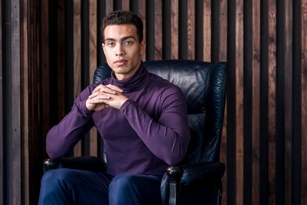 Free photo confident young man with his hand clasped sitting on cozy armchair against wooden wall