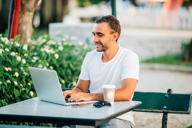 Уверенный молодой человек, работающий на ноутбуке, сидя за деревянным столом на открытом воздухе