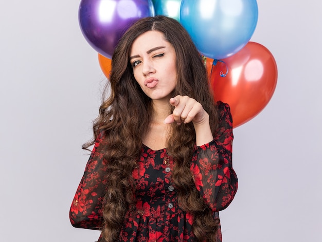 Confident young party girl holding balloons behind back looking and pointing winking isolated on white wall