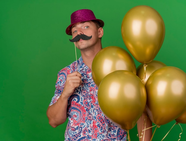 Confident young party guy wearing pink hat holding balloons putting fake mustache on stick isolated on green