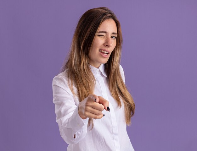 Confident young pretty caucasian girl blinks eye and points at camera isolated on purple wall with copy space