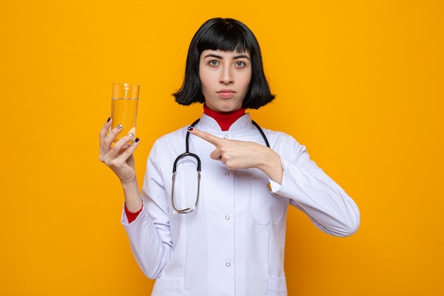 Confident young pretty caucasian girl in doctor uniform with stethoscope holding glass of water 