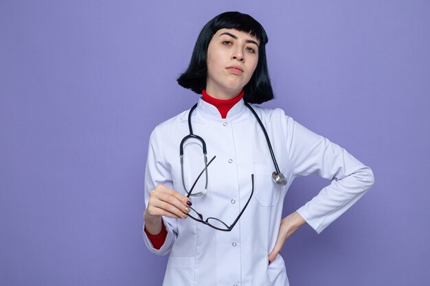 Confident young pretty caucasian girl in doctor uniform with stethoscope holding optical glasses and 