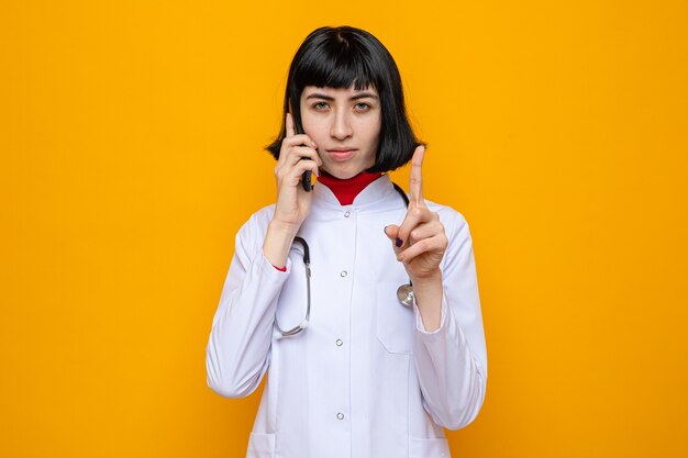 Confident young pretty caucasian girl in doctor uniform with stethoscope talking on phone and pointing up