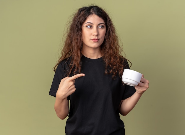 Бесплатное фото Уверенная молодая красивая кавказская девушка, держащая и указывающая на чашку чая, смотрящую в сторону