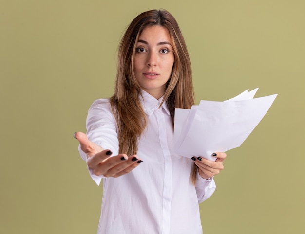 Confident young pretty caucasian girl holding blank paper sheets and stretching out hand isolated on olive green wall with copy space