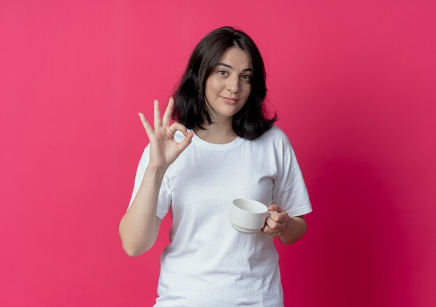 Confident young pretty caucasian girl holding cup and doing ok sign isolated on crimson background with copy space