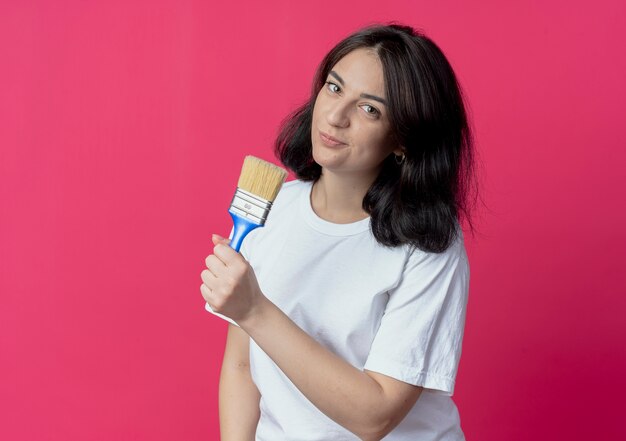 Confident young pretty caucasian girl holding paint brush isolated on crimson background with copy space