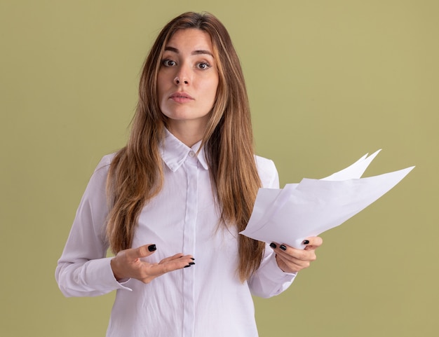 Foto gratuita la giovane ragazza abbastanza caucasica sicura tiene e indica i fogli di carta in bianco con la mano isolata sulla parete verde oliva con lo spazio della copia