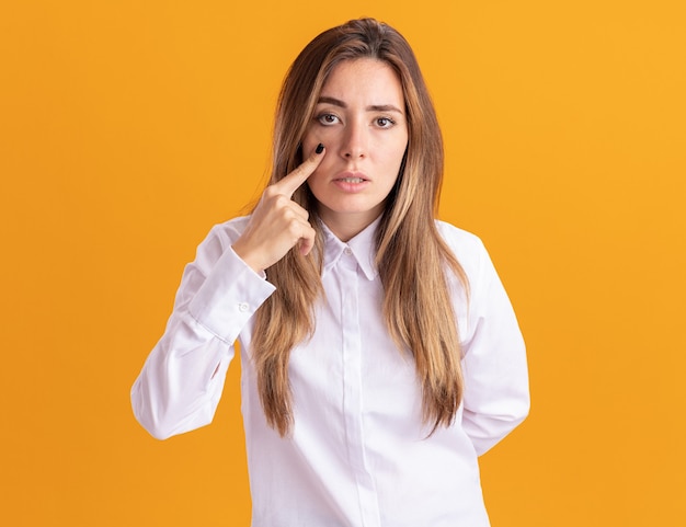 Confident young pretty caucasian girl puts finger on eyelid and looks at camera on orange 